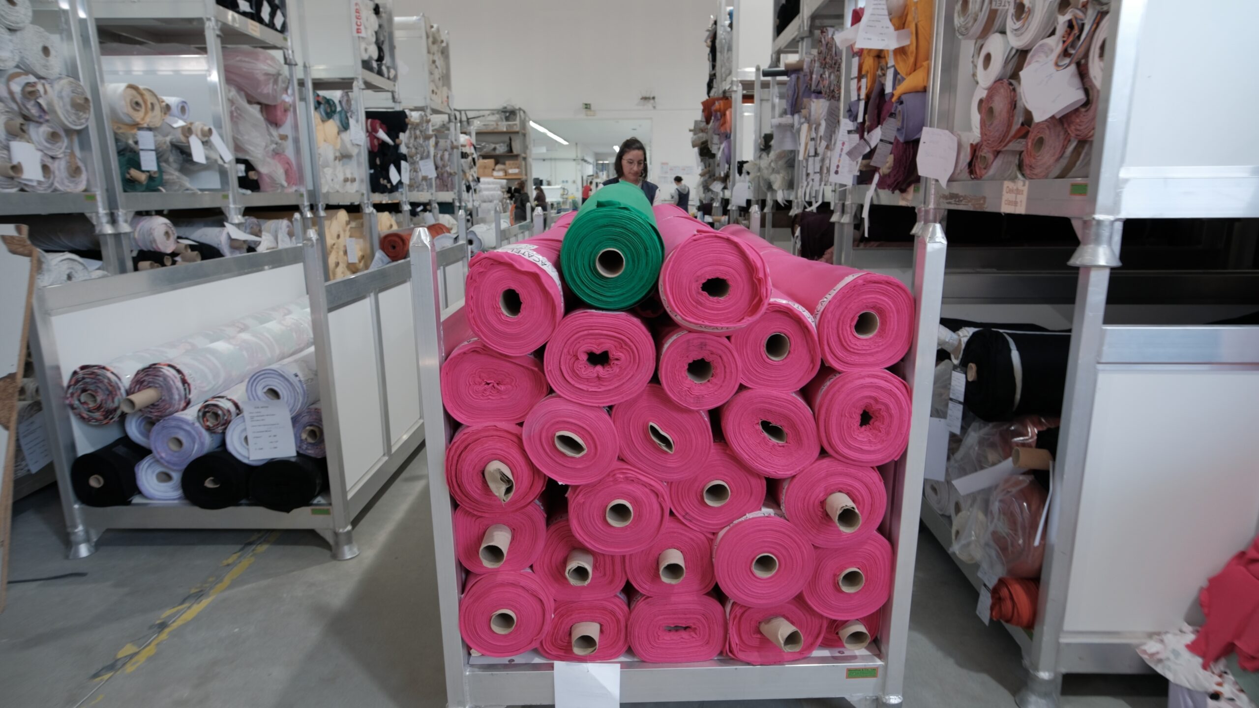 25 isoa pinkkiä neulosrullaa kasassa liikuteltavan lavan päällä Black Moda Portugal -tehtaan varastoosa. lavan takana seisoo työntekijä keskittyneenä lavan liikutteluun. Sivuilla nousevat korkeat hyllyt, joissa lisää isoja neulosrullia. 