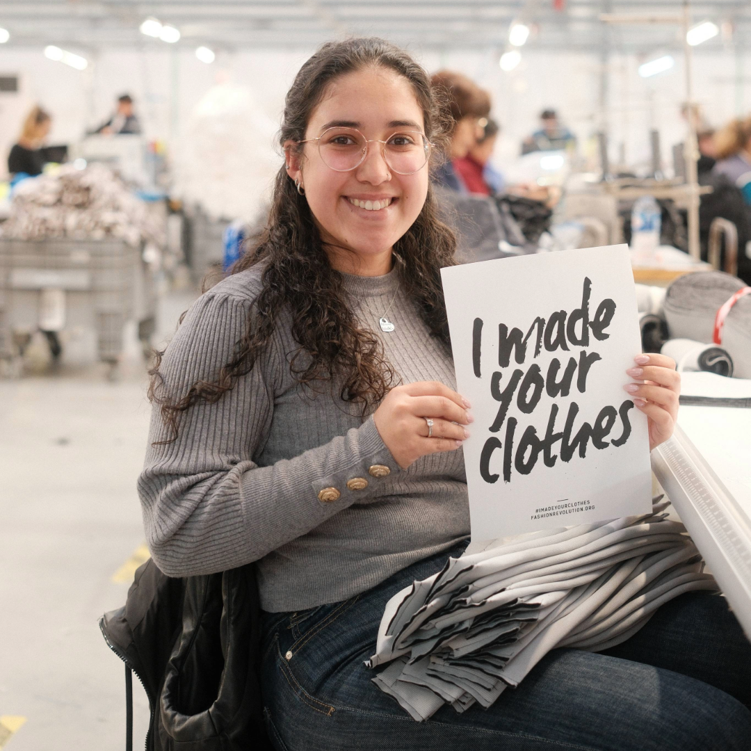 Black Moda Portugal Lda -tehtaan työntekijä istuu pöydän ääressä hymyillen ja pitäen käsissään A4-kokoista valkoista paperia, jossa lukee "I made your clothes" 