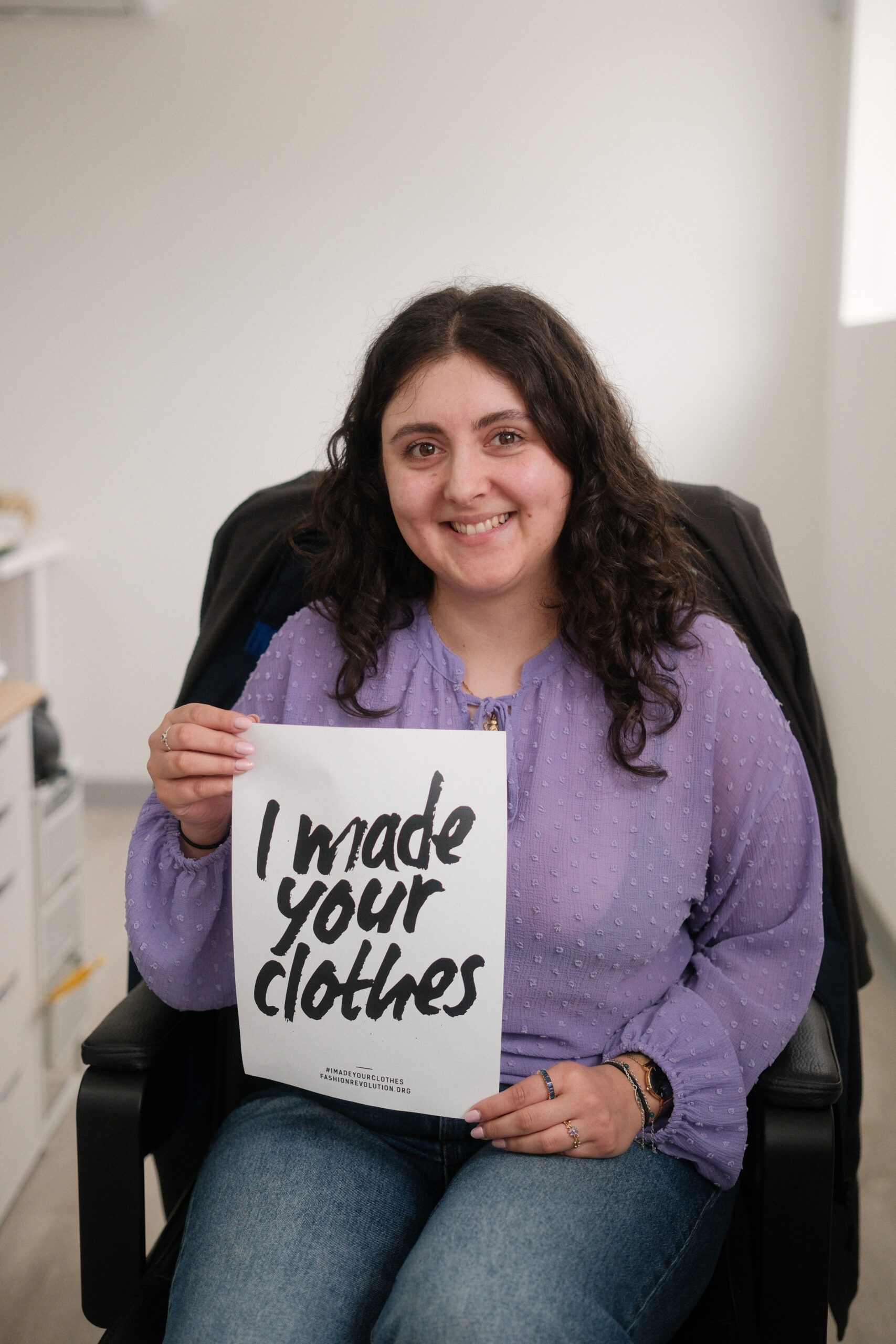 Black Moda Portugal Lda -tehtaan työntekijä istuu toimistotuolilla hymyillen ja pitäen käsissään A4-kokoista valkoista paperia, jossa lukee "I made your clothes" 