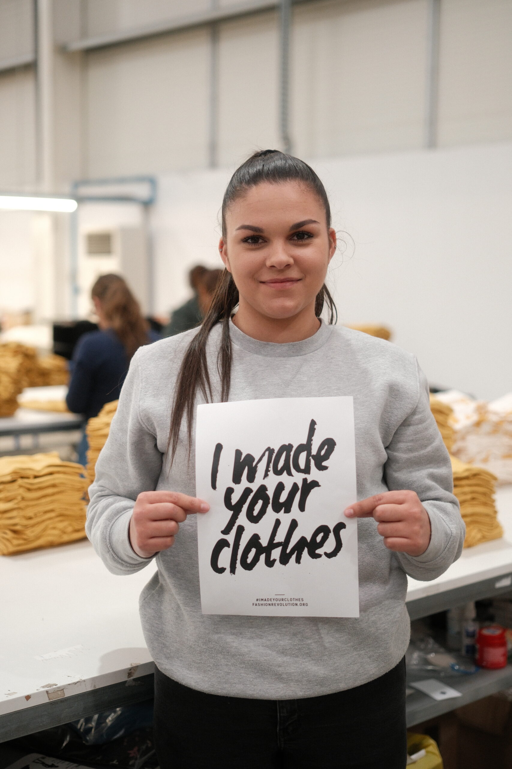 Black Moda Portugal Lda -tehtaan työntekijä seisoo pöydän edessä hymyillen ja pitäen käsissään A4-kokoista valkoista paperia, jossa lukee "I made your clothes". Takana pöydällä on keltaisia trikoovaatteita siististi pinoissa. 