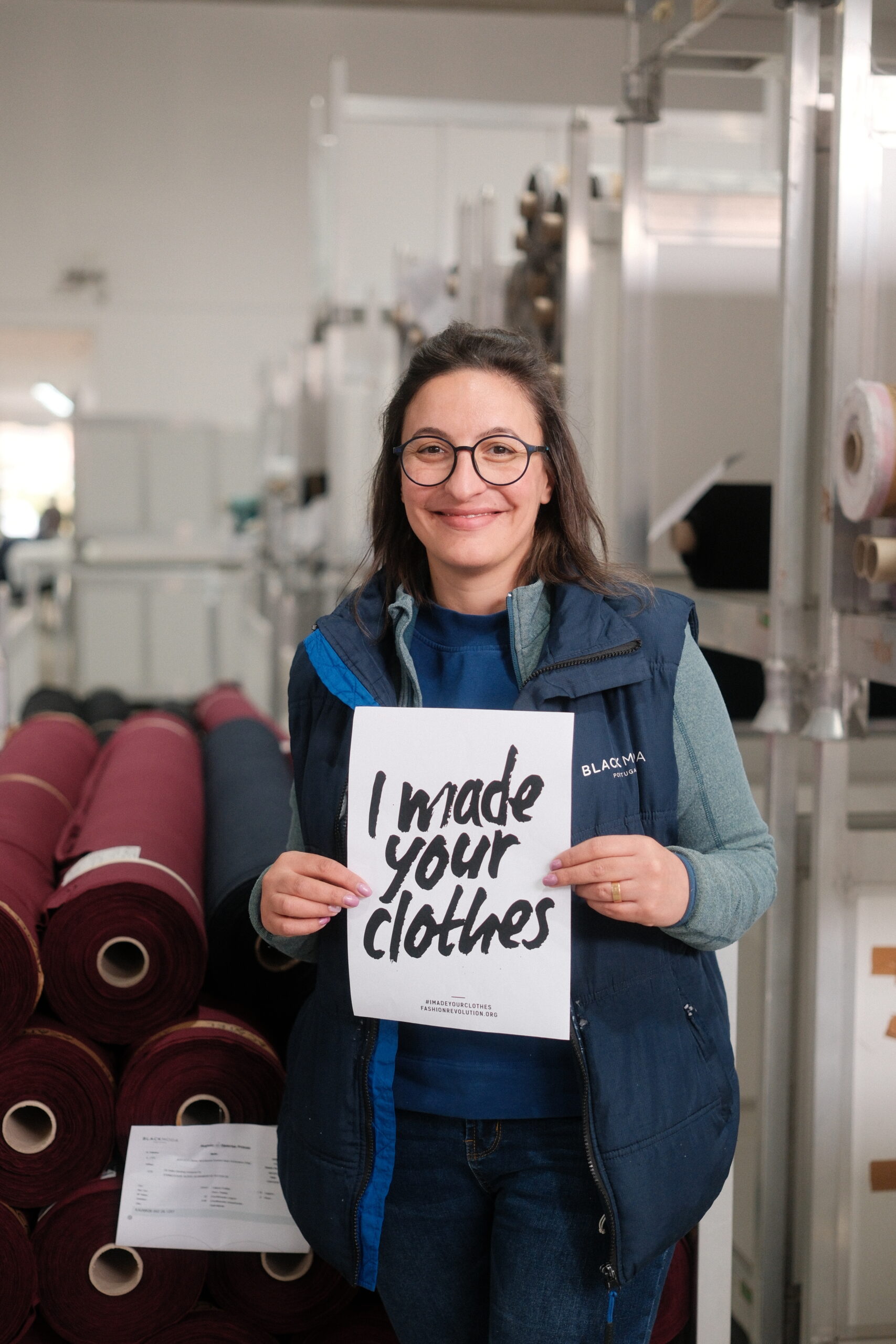 Black Moda Portugal Lda -tehtaan työntekijä seisoo isojen, viininpunaisten neulosrullien edessä hymyillen ja pitäen käsissään A4-kokoista valkoista paperia, jossa lukee "I made your clothes". 