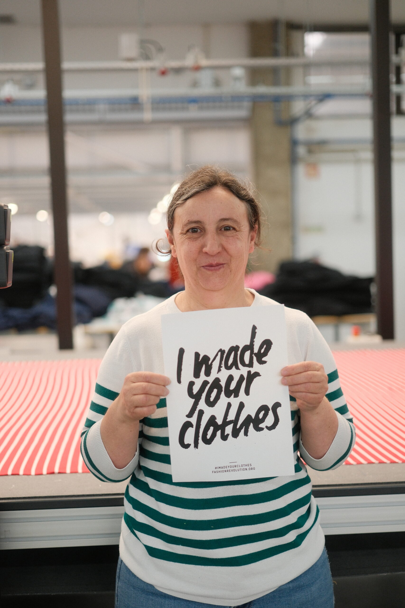 Black Moda Portugal Lda -tehtaan työntekijä seisoo leikkuupöydän edessä hymyillen ja pitäen käsissään A4-kokoista valkoista paperia, jossa lukee "I made your clothes". Leikkuupöydällä on puna-valkoraidallinen neulos levitettynä. 