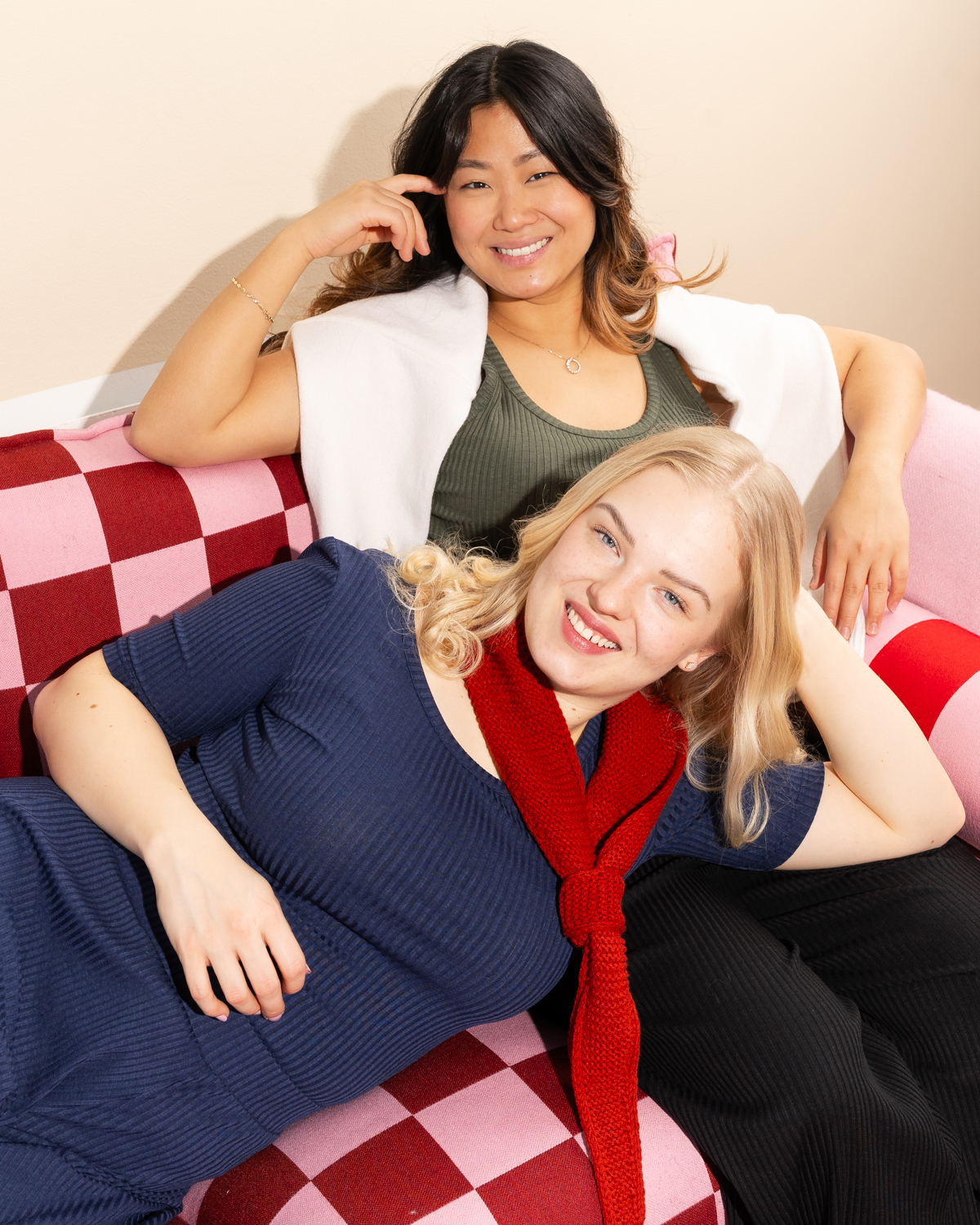 Kaksi naista loikoilevat iloisesti Toive-vaatemerkin ribbivaatteissa. Toisella on päällä sininen t-paita ja housut, toisella on päällä vihreä t-paita ja housut. Sohva on ruudullinen ja sen ruudut ovat pinkkejä ja punaisia.