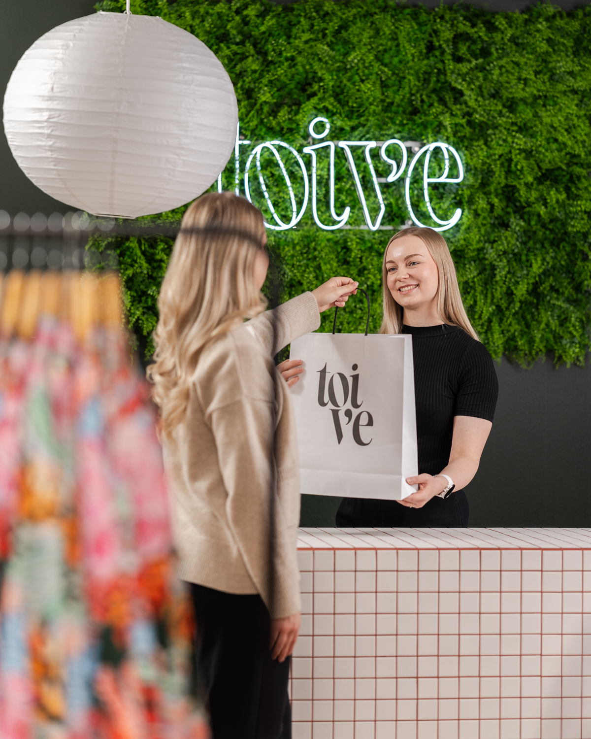 Toive-vaatemerkin liikkeeesä myyjä ojentaa kassalla asiakkaalle Toiveen logolla varustetun paperisen ostoskassin. Myyjän takana, kassanpöydän takana on kaunis viherseinä ja Toiveen logo tehtynä hohtavana neon-valona.