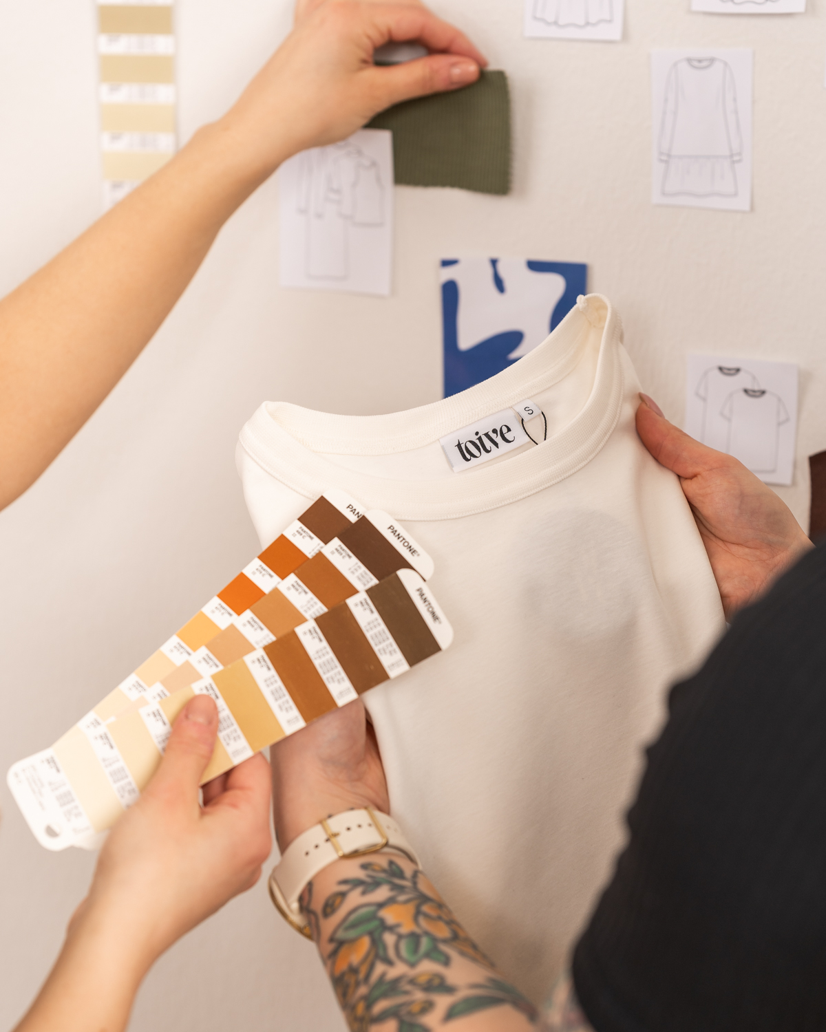 Toive-vaatemerkin toinen suuunnittelija pitää kädessään Toive-vaatemerkin kermanvaaleaa t-paitaa. Toinen suunnittelija pitää paidan edessä ruskeansävyistä värikarttaa ja toinen käsi pitelee seinällä olevaa materiaalinäytettä.