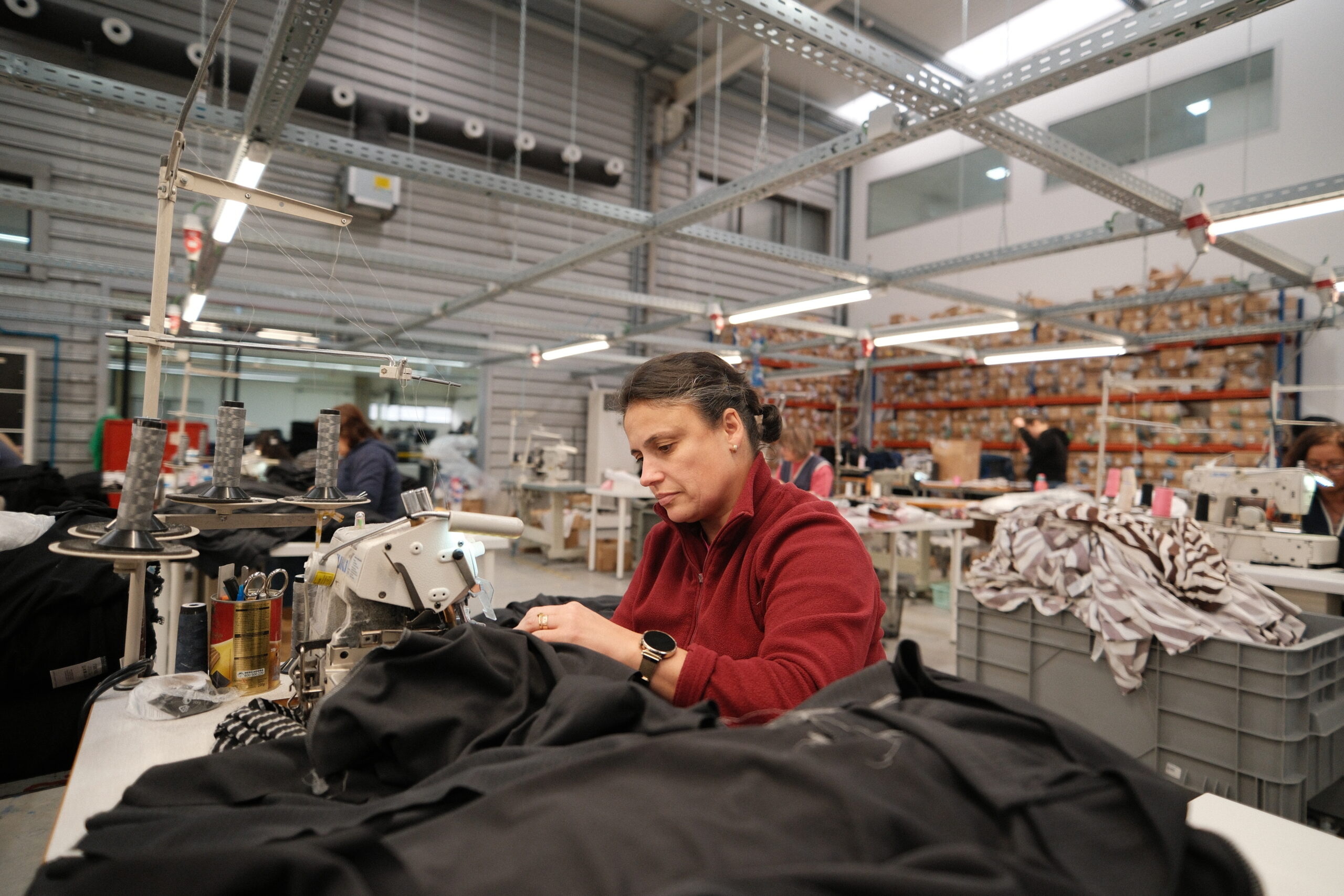 Black Moda Portugal-tehtaan työntekijä istuu teollisuussaumurin ääressä ja ompelee mustaa trikoovaatetta. Taustalla on muita työntekijöitä saumurien ääressä. 